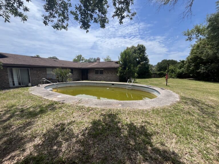 Backyard with an empty, algae-filled swimming pool and surrounding trees in Jacksonville, FL.